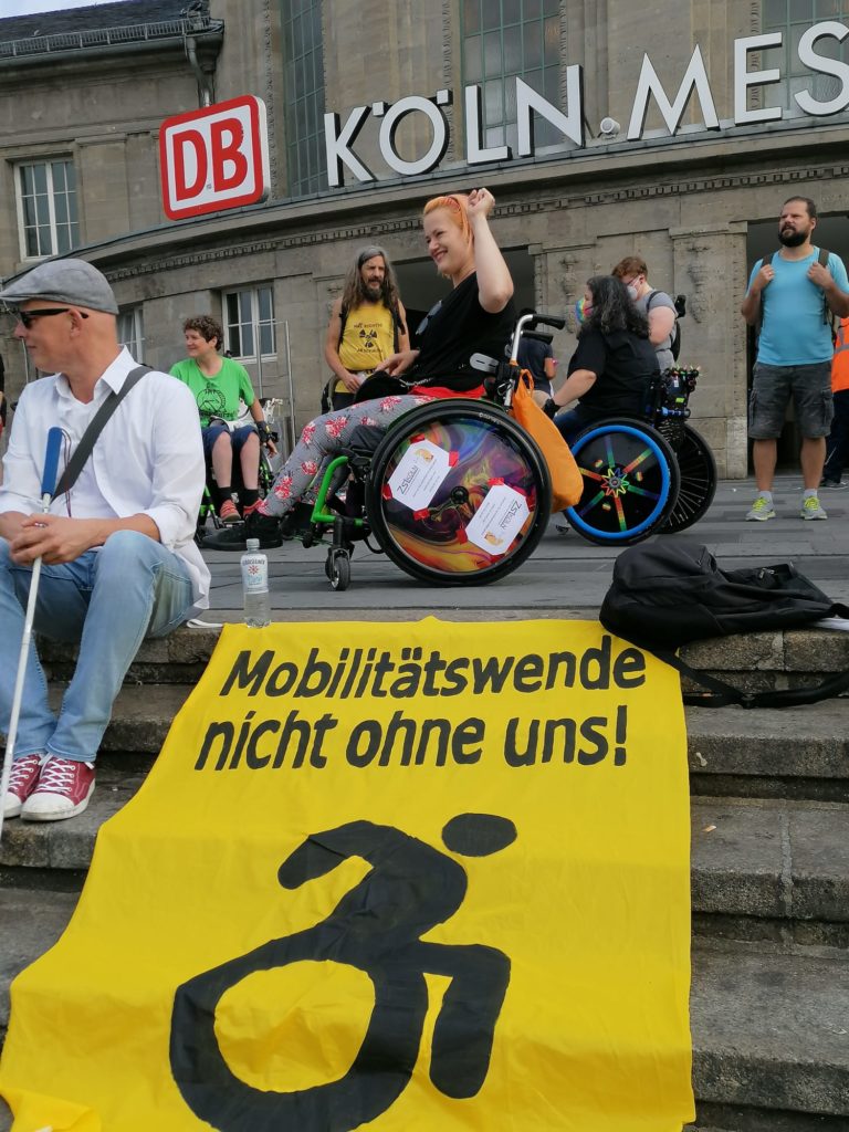 Menschen mit verschiedenen Behinderung demonstrieren auf dem Bahnhofsvorplatz. Auf den Stufen ist ein Plakat mit der Aufschrift "Mobilitätswende nicht ohne uns!" ausgebreitet.