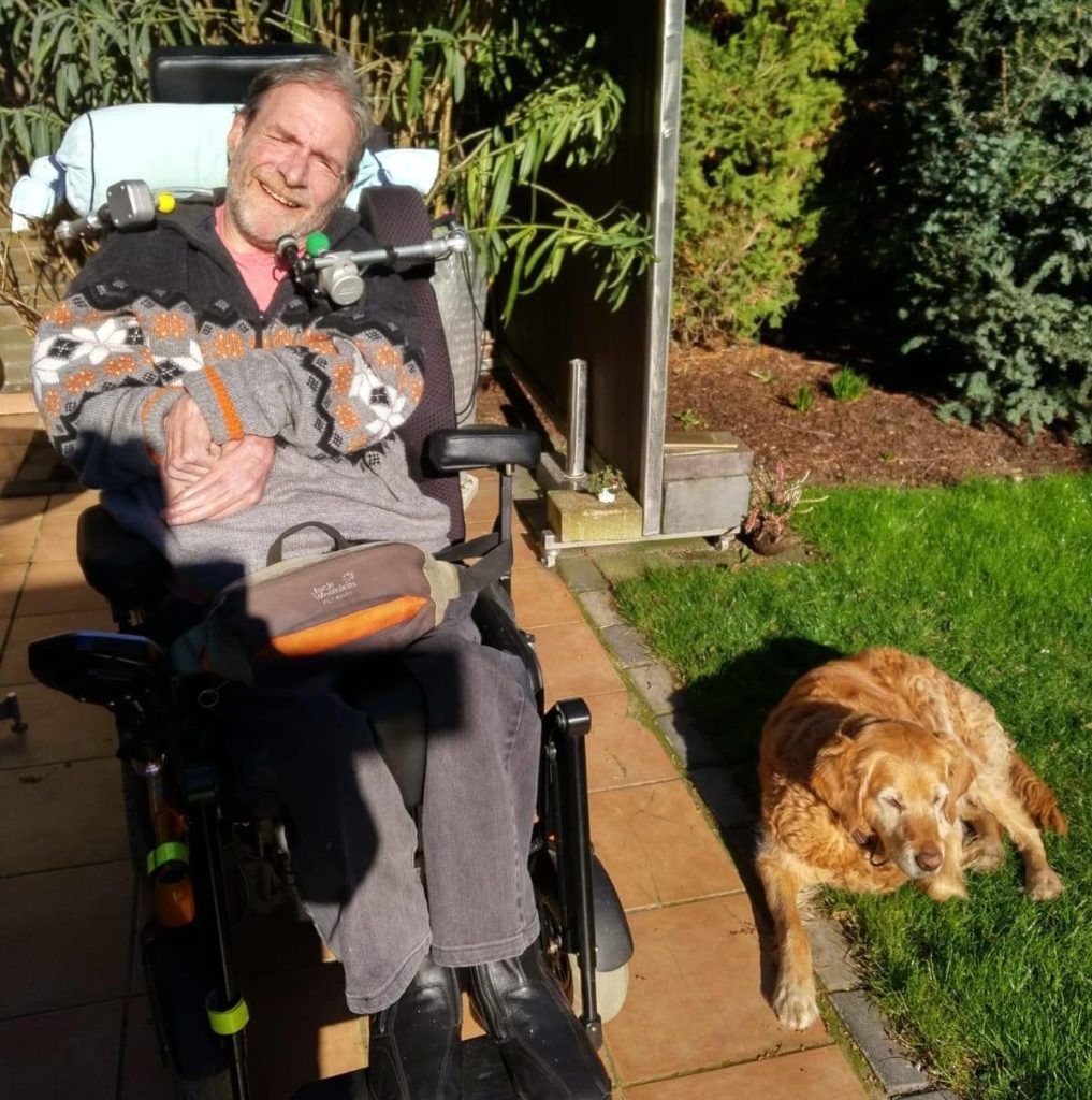 Michaels Knuffmann sitz in seinem Rollstuhl im Garten. Neben ihm liegt ein Hund im Gras.